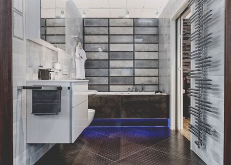 Moderne Badeausstellung mit eingelassener Badewanne, grauen Wandfliesen, beleuchtetem Spiegel und glänzendem Chrom-Heizkörper.