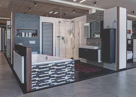 Ausstellungsbad mit halbhoher Wanne, begehbarer Dusche, Mosaikfliesen, dunklem Waschtisch und großem Spiegel.