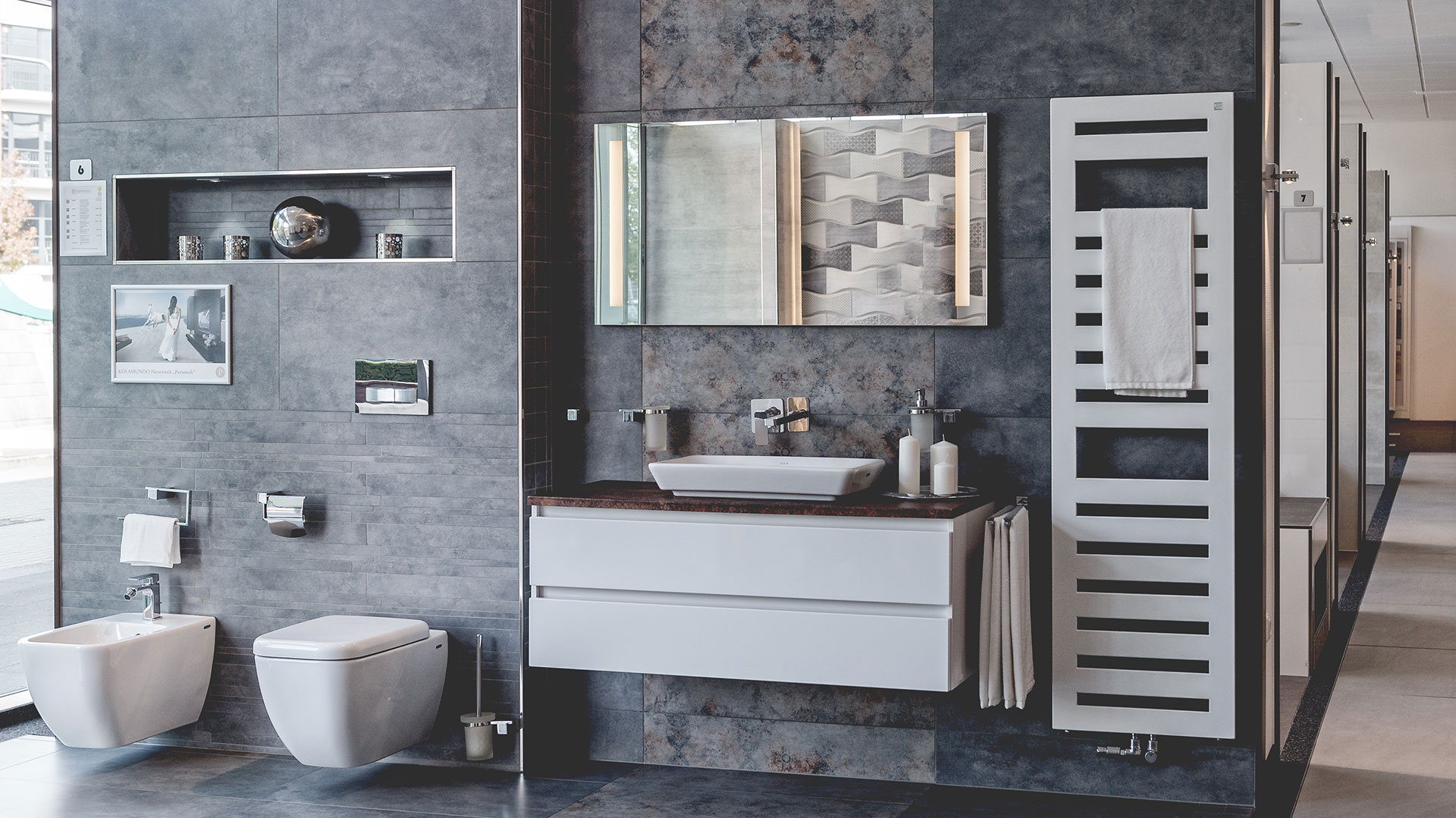 Ausstellung eines modernen Badezimmers mit grauen Wandfliesen, wandhängendem WC, Bidet, weißem Waschtisch mit dunkler Platte und beleuchtetem Spiegel.